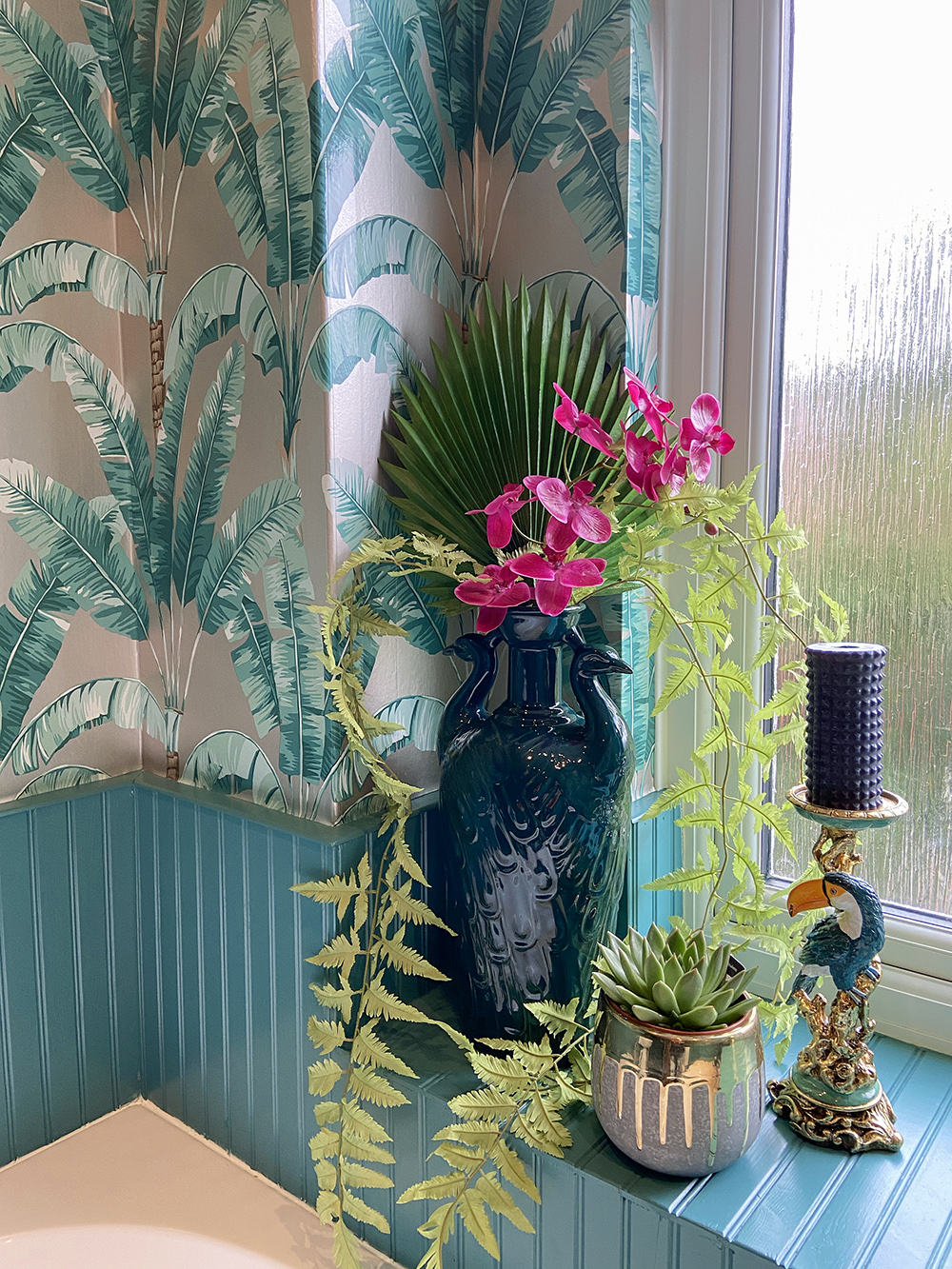 Tropical patterned wallpaper with artificial flowers and toucan candle holder to give a jungle feel